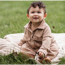 Infant wearing Burts Bees acid wash ginger loaf jumpsuit, smiling while sitting on a blanket in grass.