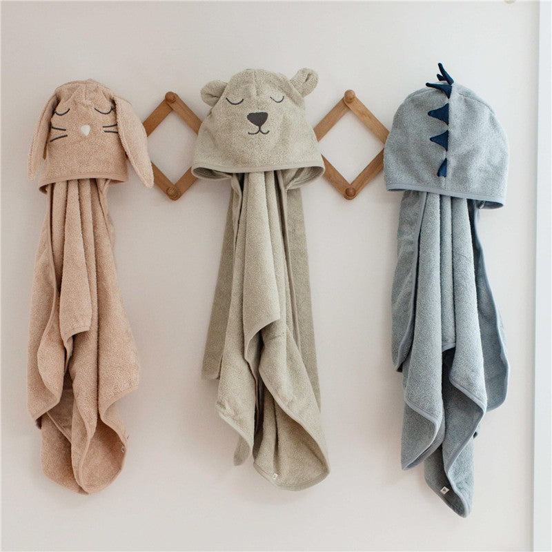 Hooded towels hanging on a wall: a bunny design in blush, a bear in taupe, and a dinosaur in blue.