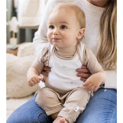 Baby wearing a beige outfit with a white bib, seated on a lap, smiling, in a cozy, neutral-toned setting.