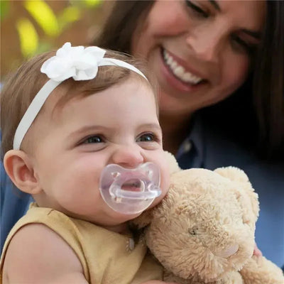 Baby smiling with a Chicco PhysioForma clear silicone pacifier, holding a stuffed bear, with a woman in the background.