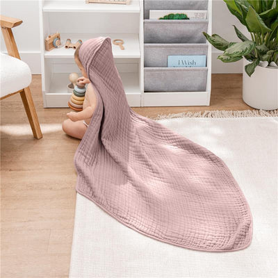 A baby seated on a light rug, wearing a mauve hooded towel, surrounded by toys and a bookshelf.