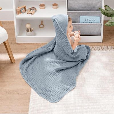 A baby wrapped in a soft Pacific blue hooded towel, sitting on a light rug, holding a toy, with shelves in the background.