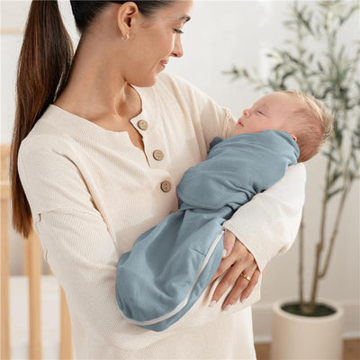 Woman holding a swaddled baby in a light blue Comfy Cubs swaddle; cozy indoor setting with greenery in the background.