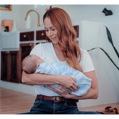 Mother gently holds a wrapped baby in blue and white striped swaddle, smiling in a cozy indoor setting.