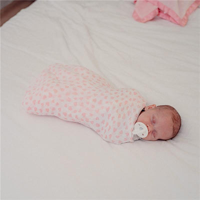 A sleeping baby swaddled in a light pink muslin blanket with a heart pattern, using a pacifier, on a white bed.