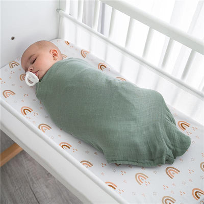 Sleeping baby swaddled in a sage green muslin blanket, resting on a crib sheet with rainbow patterns.