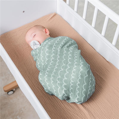 Baby swaddled in a green muslin blanket with white waves, resting peacefully in a crib with a tan mattress.