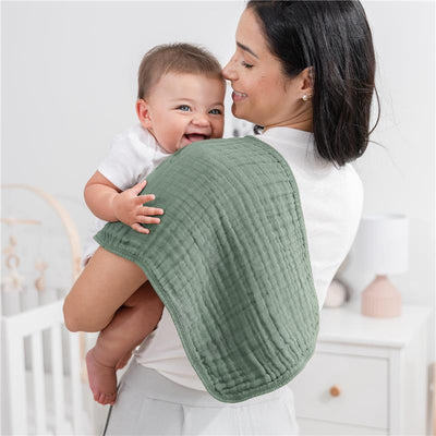 Mom holds a smiling baby over her shoulder, using a soft green muslin burp cloth. The background features a nursery.