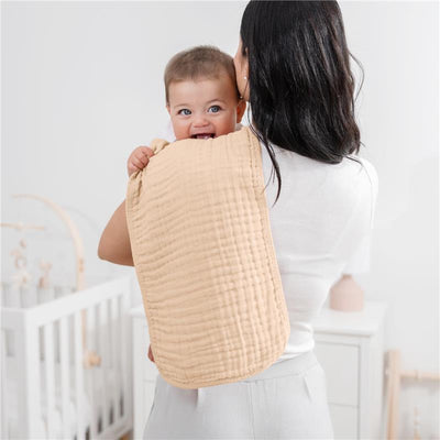 A woman holds a smiling baby against her shoulder, using a soft, beige muslin burp cloth for feeding.
