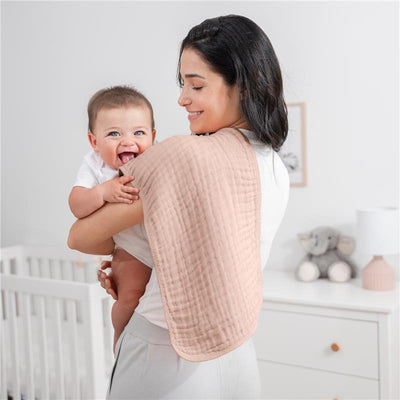 Mom smiles while holding a baby, wearing a blush muslin burp cloth draped over her shoulder, in a nursery setting.