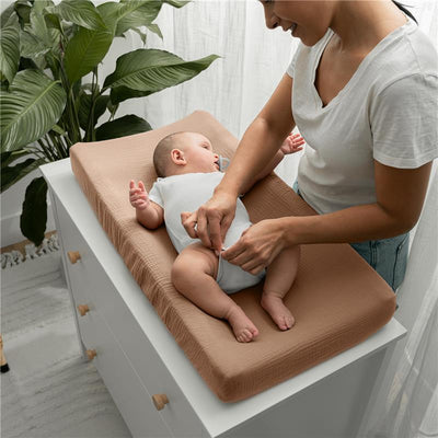 Muslin fitted changing pad cover in cedar color on a changing table with a parent gently dressing a baby.