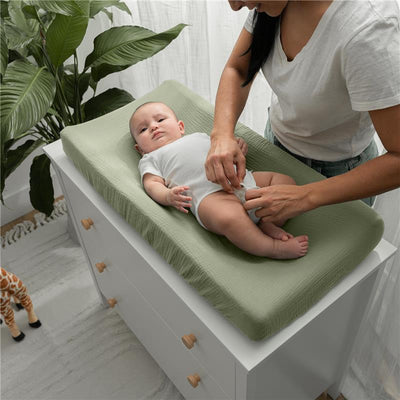 Muslin fitted changing pad cover in sage on a dresser, with a caregiver changing the diaper of a baby.