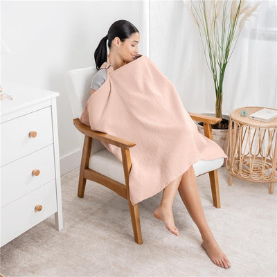 A woman seated in a chair uses a blush-colored muslin nursing cover, creating a cozy atmosphere.