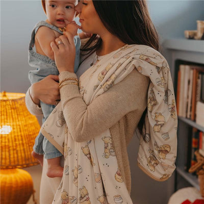 Mother holds a baby while draped in a large, stretchy quilt blanket with a playful Winnie the Pooh design.