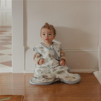 Baby sitting on a wooden floor wearing a Copper Pearl 3-Layer Knit Sleep Bag with whimsical prints and a bow headband.
