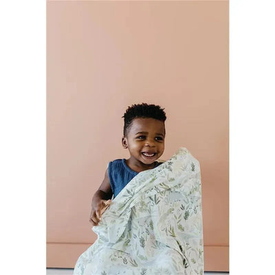 Young child smiling while holding a Copper Pearl Rex Knit Baby Swaddle Receiving Blanket with a leafy pattern.