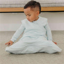 Toddler wearing a light blue Copper Pearl sleep bag, seated on a carpet, looking down curiously.