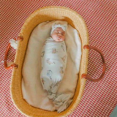 A wrapped baby sleeping peacefully in a woven bassinette, covered with a Copper Pearl Winnie the Pooh knit blanket.