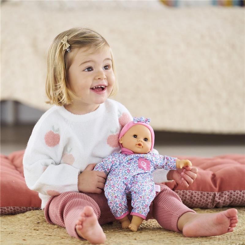 Smiling child in a white sweater holds a Corolle Bébé Calin Capucine doll, wearing floral pajamas and a pink headband.