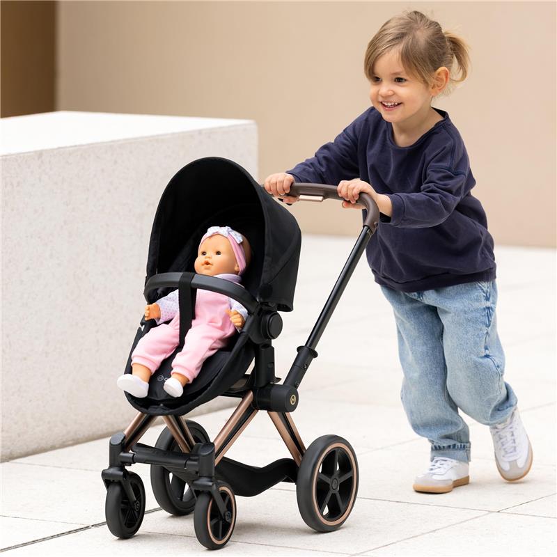Young child pushing a Corolle Dolls stroller, featuring a black frame and a doll dressed in pink seated inside.