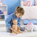 Young girl plays with a Corolle doll, sitting on a rug with a changing accessory set, including a bag and doll items.