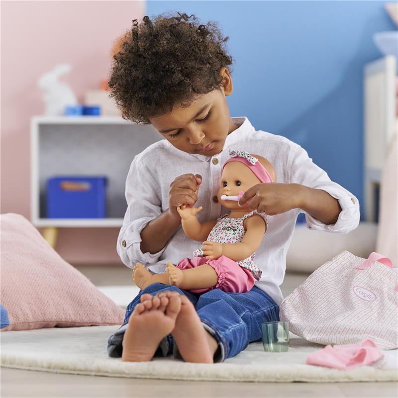 A child in a white shirt is grooming a Corolle doll wearing a floral outfit, with a pink headband, using a toothbrush.