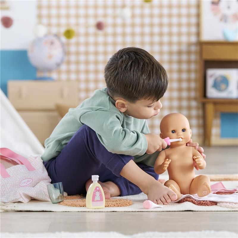 Child using a toothbrush on a Corolle doll, with accessories including a pink bag, lotion bottle, and glass.