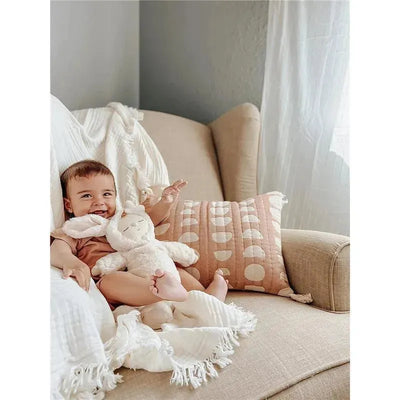 Baby sits on a beige chair with a plush bunny toy, surrounded by soft blankets and a textured pink moon phase pillow.