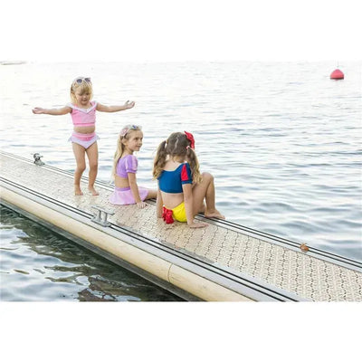 Three children in colorful swim suits play on a dock by the water, with one girl jumping joyfully.