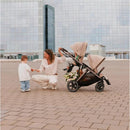 Cybex Gazelle S Second Seat in taupe and chocolate brown, with a woman interacting with a child beside the stroller.
