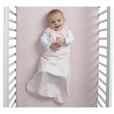 Smiling baby in a pink and white striped sleep sack with built-in swaddle, lying in a crib with a pink dotted sheet.