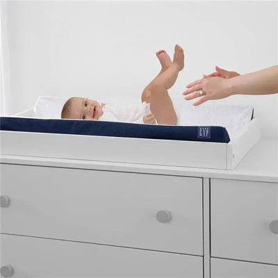 Contoured changing pad with navy cover holds a baby, while an adult hand reaches toward them, on a white dresser.