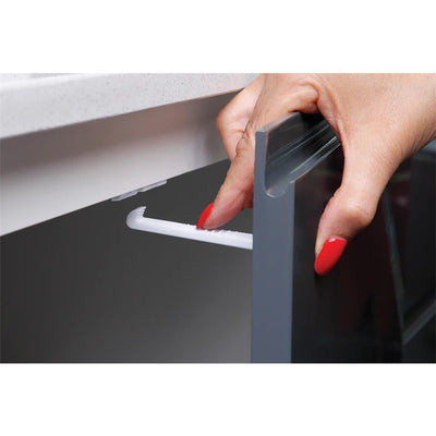 A hand with red nails is shown installing a white Dreambaby safety catch onto a cabinet door for child safety.
