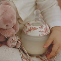 Elhee Baby Bottle with floral design held by a child, alongside a soft pink comforter and plush toy.