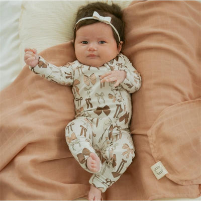 Baby wearing a beige and brown bow-patterned zipper romper, lying on a tan blanket, with a matching bow headband.