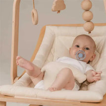 Baby in a light-colored baby seat with a pacifier, surrounded by wooden mobile toys, appearing calm and relaxed.