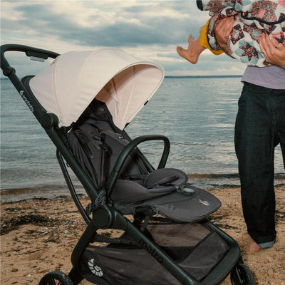 Ergobaby Metro 3 stroller on a beach, showcasing a white canopy, adjustable seat, and a parent holding a child nearby.