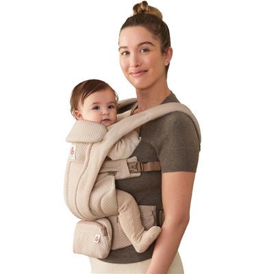 Ergobaby Omni Deluxe Baby Carrier in cream, worn by a woman holding a baby facing forward, features padded straps and adjustable buckles.