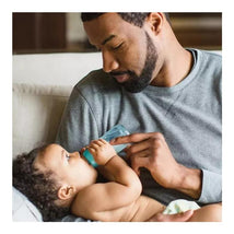 A father feeds a baby with a turquoise Evenflo Feeding plastic bottle while sitting on a couch.