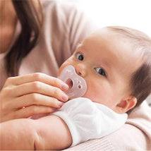 A baby holds a clear Evenflo Feeding Balance+ pacifier, while a caregiver gently supports them.