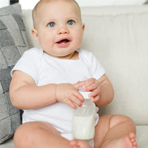 Baby sitting on a sofa, smiling while holding an 8 oz Evenflo glass bottle filled with milk.