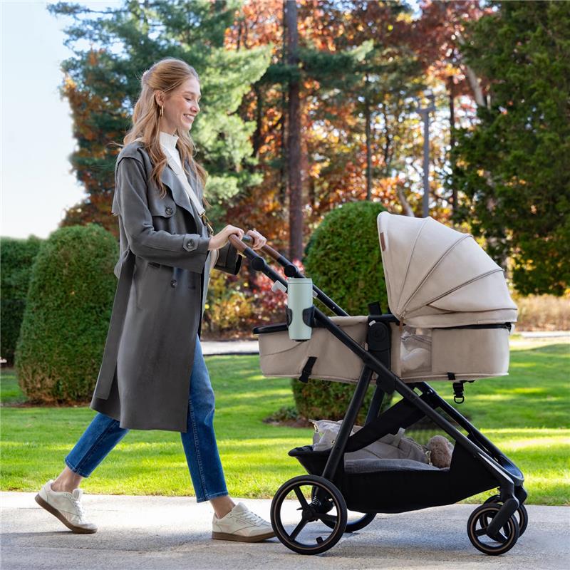 Woman walking with Evenflo Shyft Intuiti Travel System stroller in Jasper Black, featuring a beige infant car seat and cup holder.