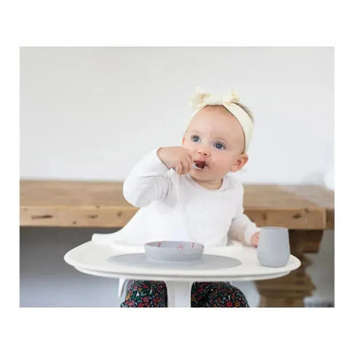 Toddler sitting in a high chair, eating from a grey bowl and holding a grey cup, wearing a white shirt and patterned leggings.