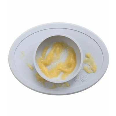 Ezpz Tiny Bowl in pewter color, featuring a curved edge, with a portion of yellow food inside and some spilled around.