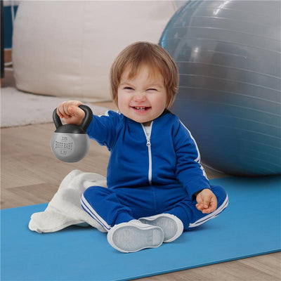 Smiling baby in a blue tracksuit holding a Buff Baby Kettlebell Rattle on a yoga mat, with an exercise ball in the background.