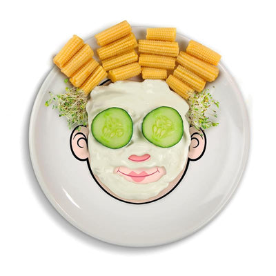 A playful dinner plate designed to resemble a face, featuring cucumbers for eyes, with corn sticks as hair and sprouts as decoration.