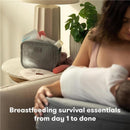Frida Mom 13Pk Breastfeeding Survival Kit in a gray bag on a table beside a nursing mother and baby, featuring various breastfeeding essentials.