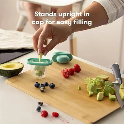 Teal Fridababy Push Pop Feeder being filled with fruit, surrounded by chopped avocado, berries, and a knife on a cutting board.