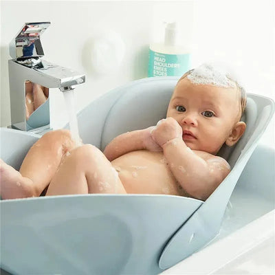 A baby sits in a soft, blue sink bath with bubbles, while water flows from a faucet nearby.