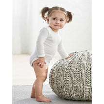 Baby wearing a white long-sleeve onesie with mitten cuffs, standing beside a knitted pouf in a bright room.